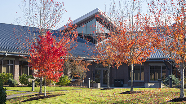 Exterior view of a modern building with a sloped roof, surrounded by vibrant red and orange autumn trees under a clear blue sky.