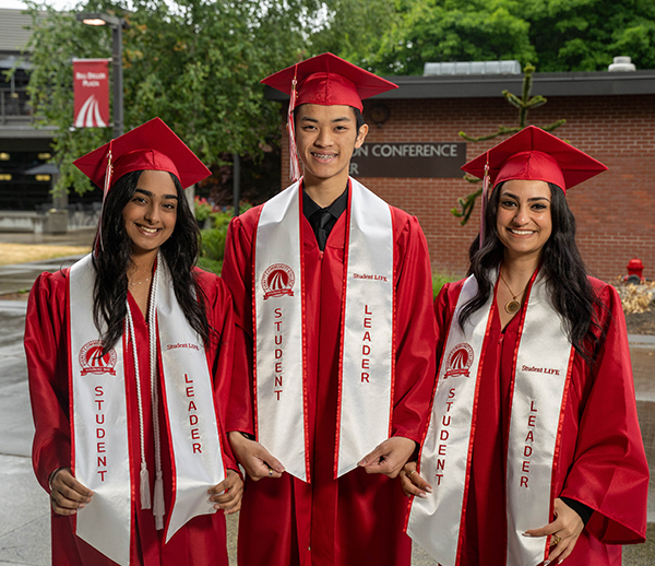 Anthony Do is centered in a photo between two class mates. They are all dressed in red graduation gowns and caps and standing on EvCC's campus with Jackson Conference Center in the background.