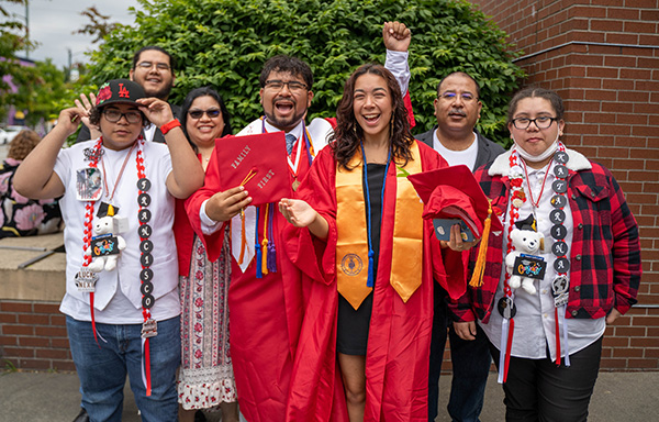 Two EvCC graduates wearing red graduation caps and gowns celebrate with members of their family.