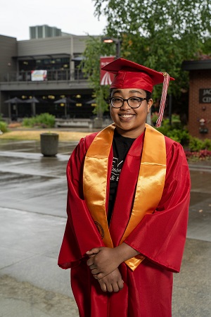 Sandra Hernandez stands in front of EvCC buildings and tall green tree, wearing red graduation cap & gown with gold stoll around her shoulders and hands clasped