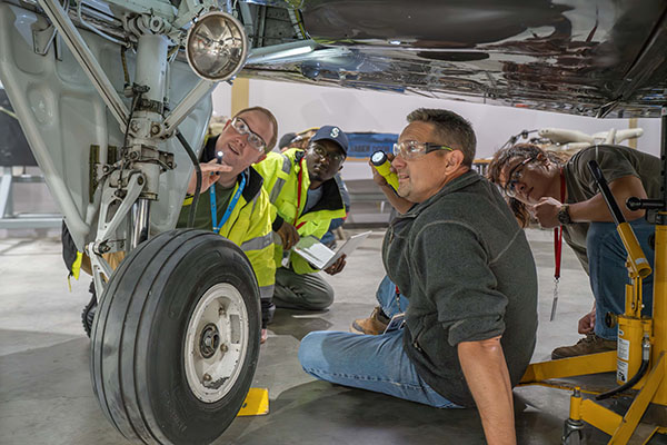 EvCC Aviation student Shad Fleshman-Cooper works on landing gear of aircraft while instructor, Christopher Russell, and fellow students look on.