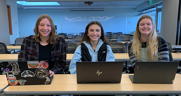 Three female students sit in a classroom behind their open laptops and smile at the camera.