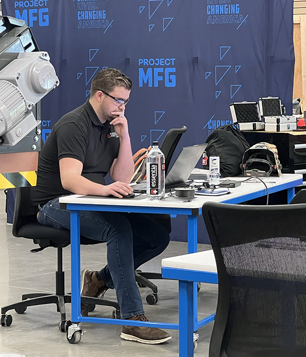 River Stormo sits at a table and works on a laptop in front of a large blue banner that says PROJECT MFG.