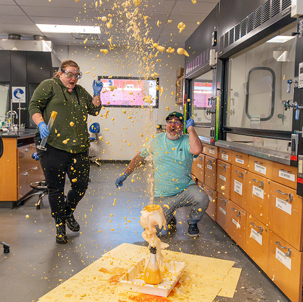 Two people wearing safety goggles look surprised and excited while yellow foam sprays out of a test tube into the air. The setting is a science lab.