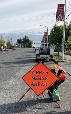 A worker sets up an orange construction zone sign that says "ZIPPER MERGE AHEAD" on the side of a road. A nearby pole has an Everett Community College banner on it.